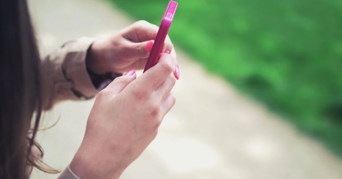 Femme tenant un smartphone rose avec les deux mains dans un cadre extérieur décontracté.
