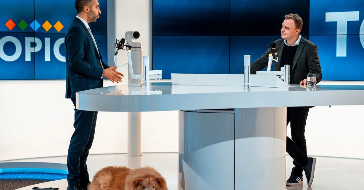 Deux animateurs discutent dans un studio de télévision moderne, un chien couché à leurs côtés. Un décor professionnel soigné.